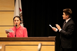 [ai] A woman in a bright pink blouse speaks at a podium while holding notes. A man in a black suit gestures as he listens attentively, with a flag visible in the background.